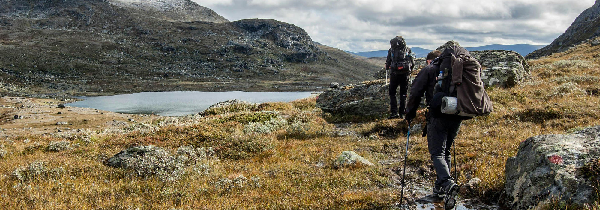 Two hikers on a mountain trail with a lake and mountains in the background
