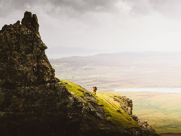 Person standing on a grassy cliff overlooking a landscape with a large rock formation.