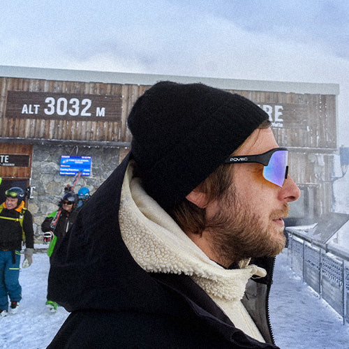 Person wearing a black beanie and sunglasses in a snowy landscape with a sign indicating altitude.