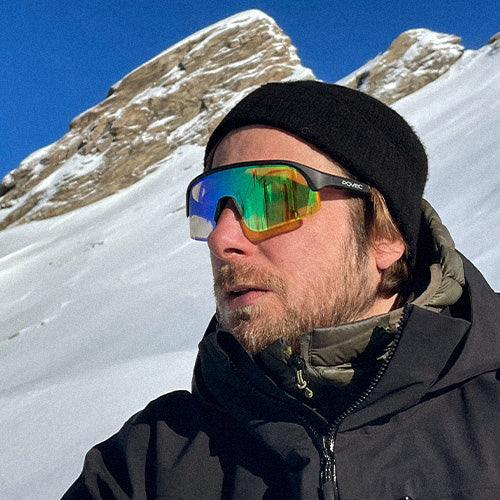 Person wearing reflective sunglasses and a black beanie with a snowy mountain background